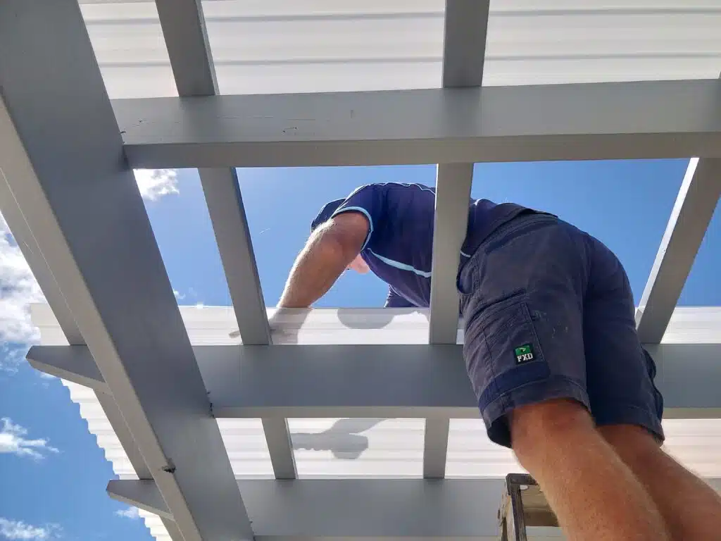 Tradesperson installing a white timber pergola structure viewed from below against a blue sky at a Two Rocks property