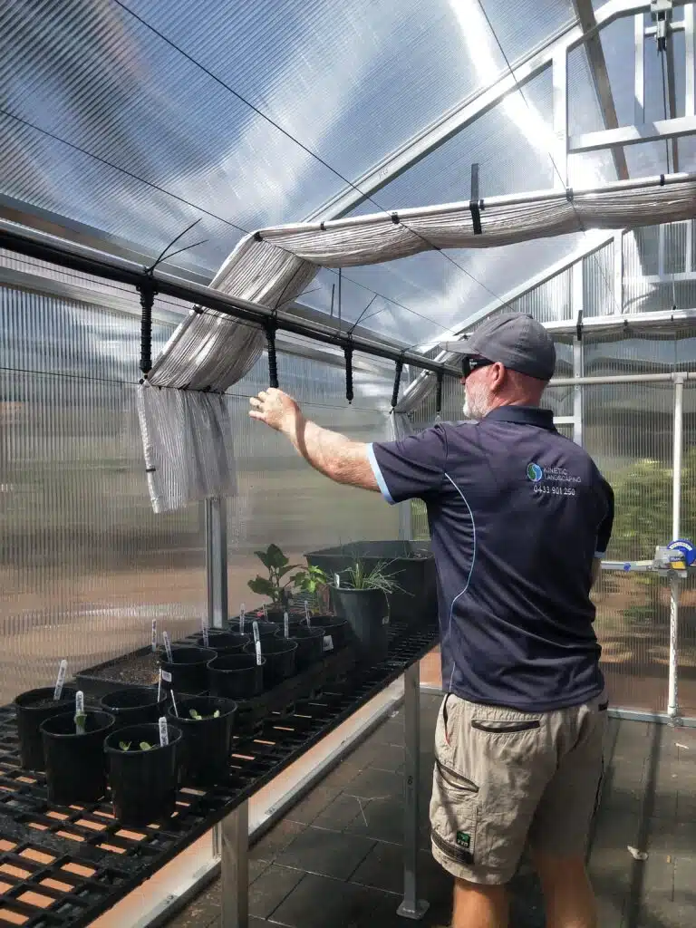 Landscaper adjusting overhead misting lines inside a clear polycarbonate greenhouse.