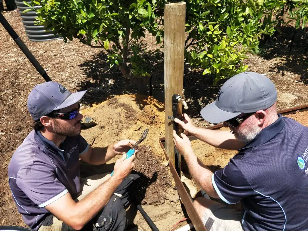 Two landscapers installing an outdoor tap and irrigation connection beside a timber post.