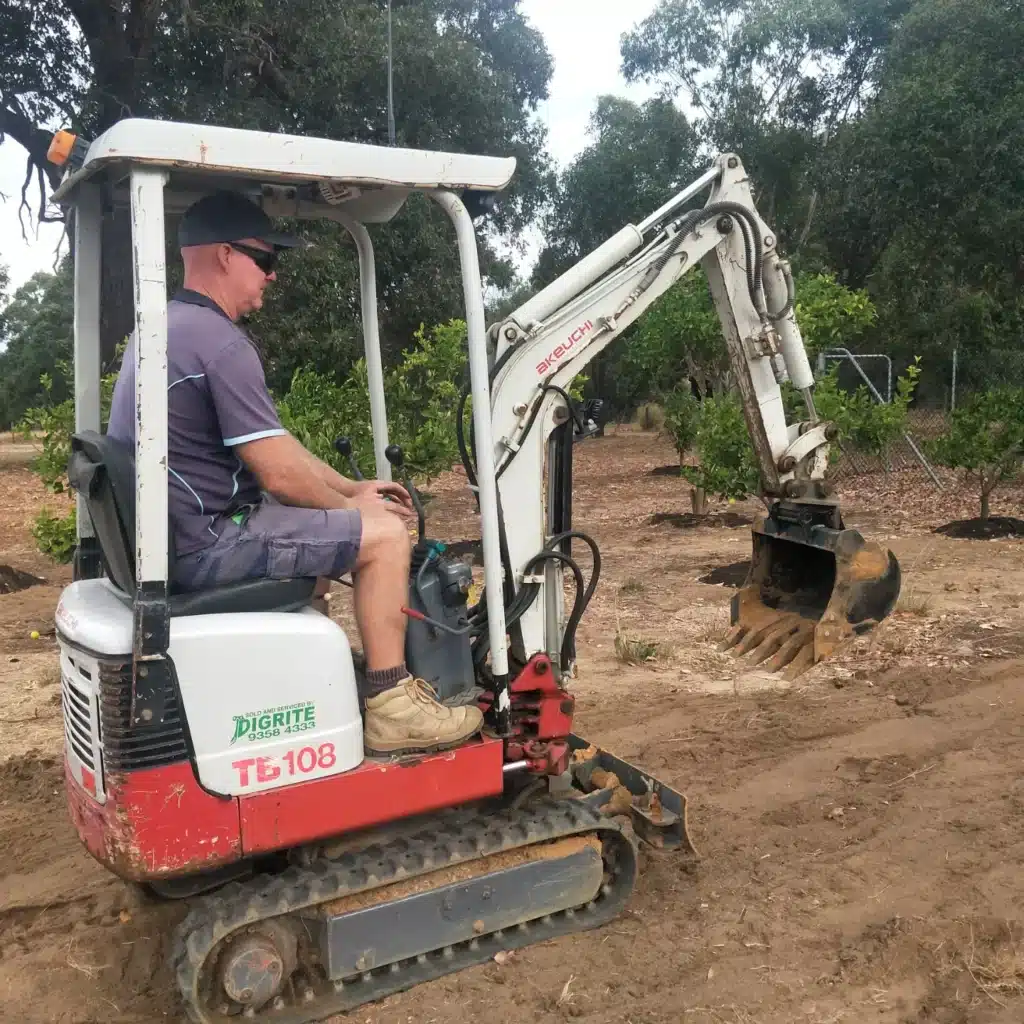 Landscaper operating a compact excavator while digging soil among orchard trees.